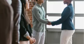 Two adults shaking hands with one congratulating another with onlookers.